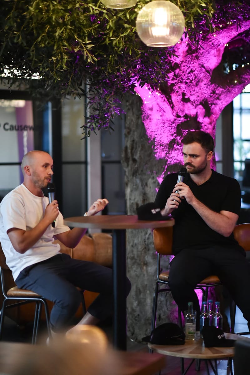 Two speakers with microphones in conversation on stage beside the lit tree at 1 Mill Street