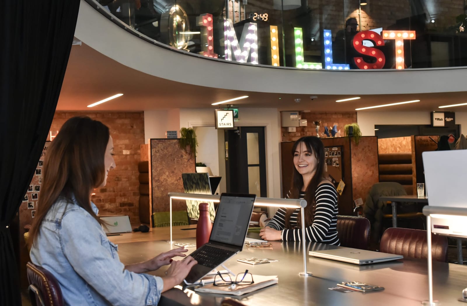 Two members working at hot desks under the Mill St light-up letters