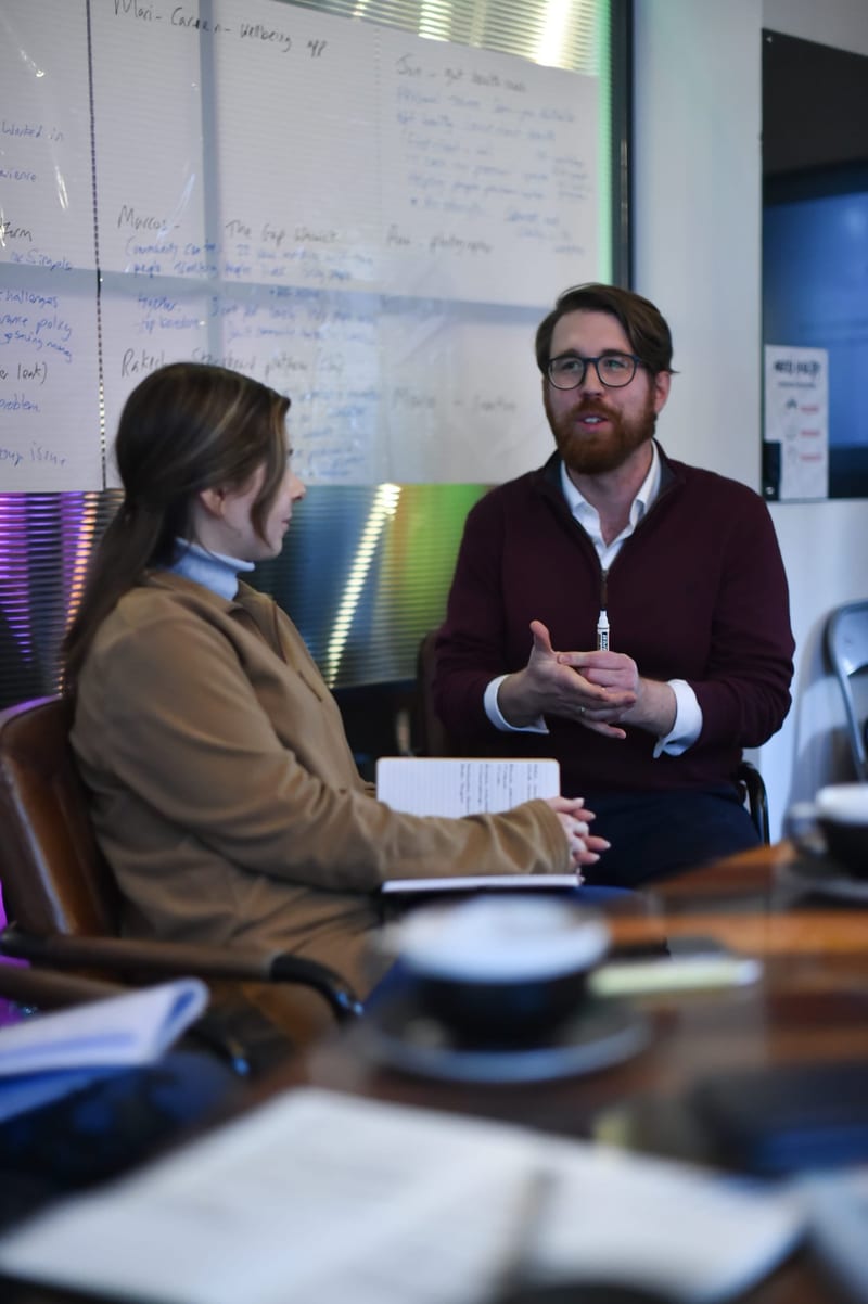 Two people talking through ideas at a table in front of a whiteboard