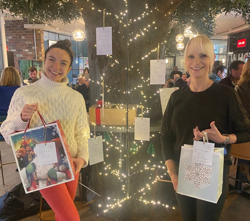 Two people holding Christmas gift bags in front of the lit-up tree at 1 Mill Street